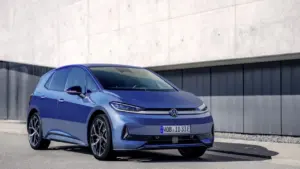 Mėlynas „Volkswagen ID.3“ elektromobilis stovi ant asfaltuoto kelio šalia modernaus betono sienos pastato.