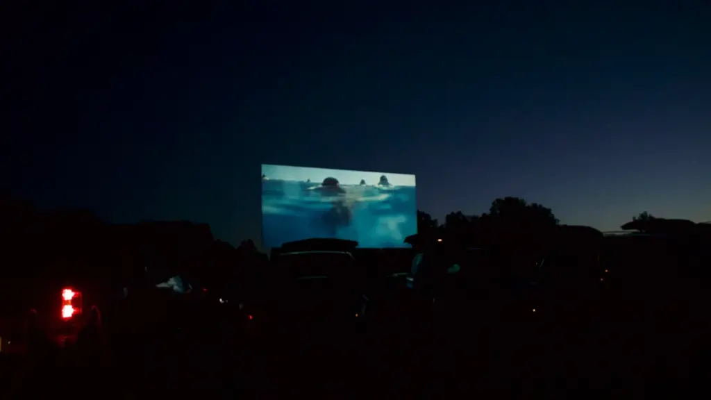 Didelis kino ekranas po atviru dangumi rodo filmą tamsiuoju paros metu lauke.