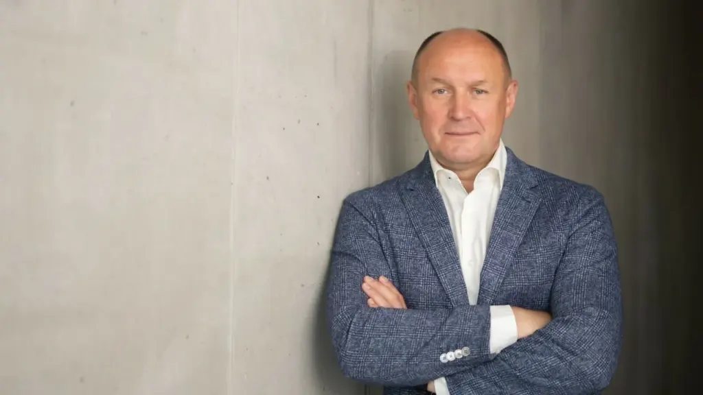 Vidutinio amžiaus vyras su šviesiu švarku ir baltais marškiniais stovi atsirėmęs į pilką sieną.