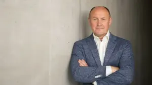 Vidutinio amžiaus vyras su šviesiu švarku ir baltais marškiniais stovi atsirėmęs į pilką sieną.