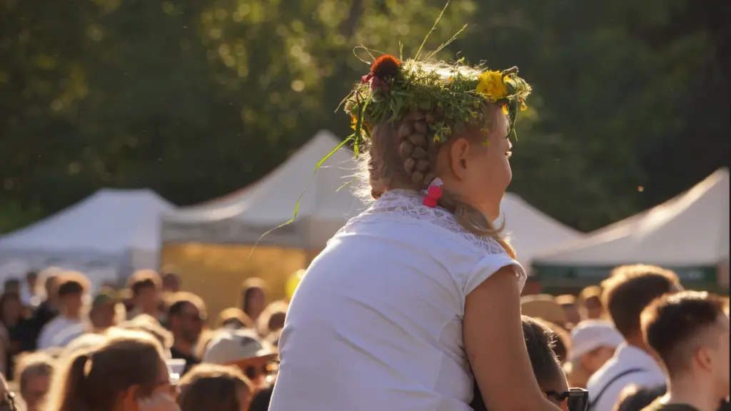Maža mergaitė su lauko gėlių vainiku ant galvos sėdi tėčiui ant pečių minioje.