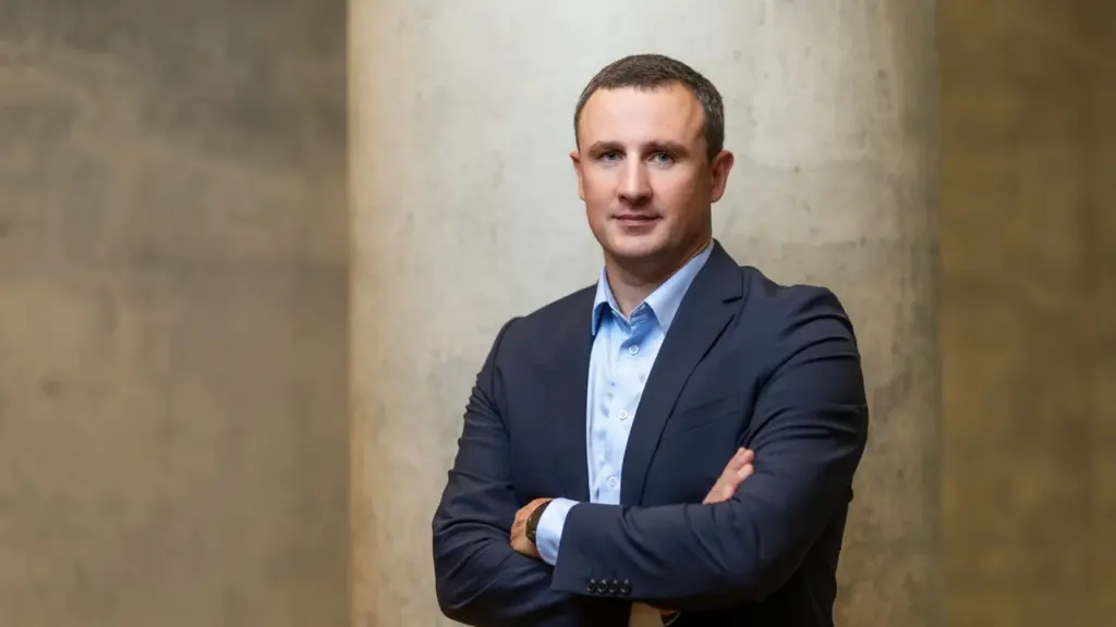 Trumpų plaukų vyras vilki tamsų švarką ir šviesius marškinius, stovi sukryžiavęs rankas ant krūtinės.