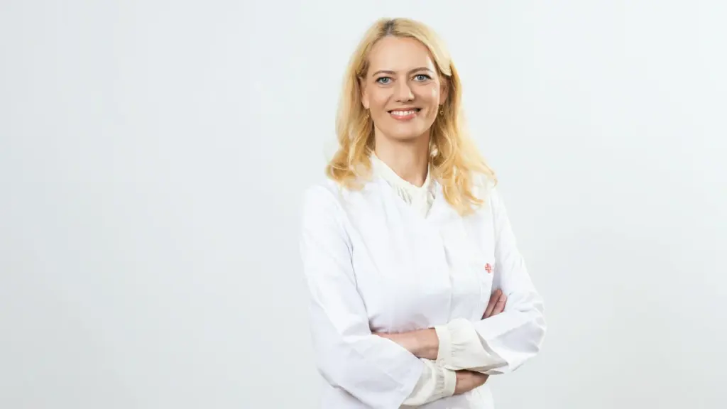 Šviesiaplaukė gydytoja su baltais medicininiais drabužiais stovi sukryžiavusi rankas ir šypsosi balto fono.